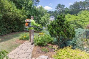 Mosquito Treatment - ceirrigation.com - ceirrigation.com Groundskeeper in a neon shirt uses a backpack sprayer to mist plants along a stone path in a sunny garden in front of a white house.